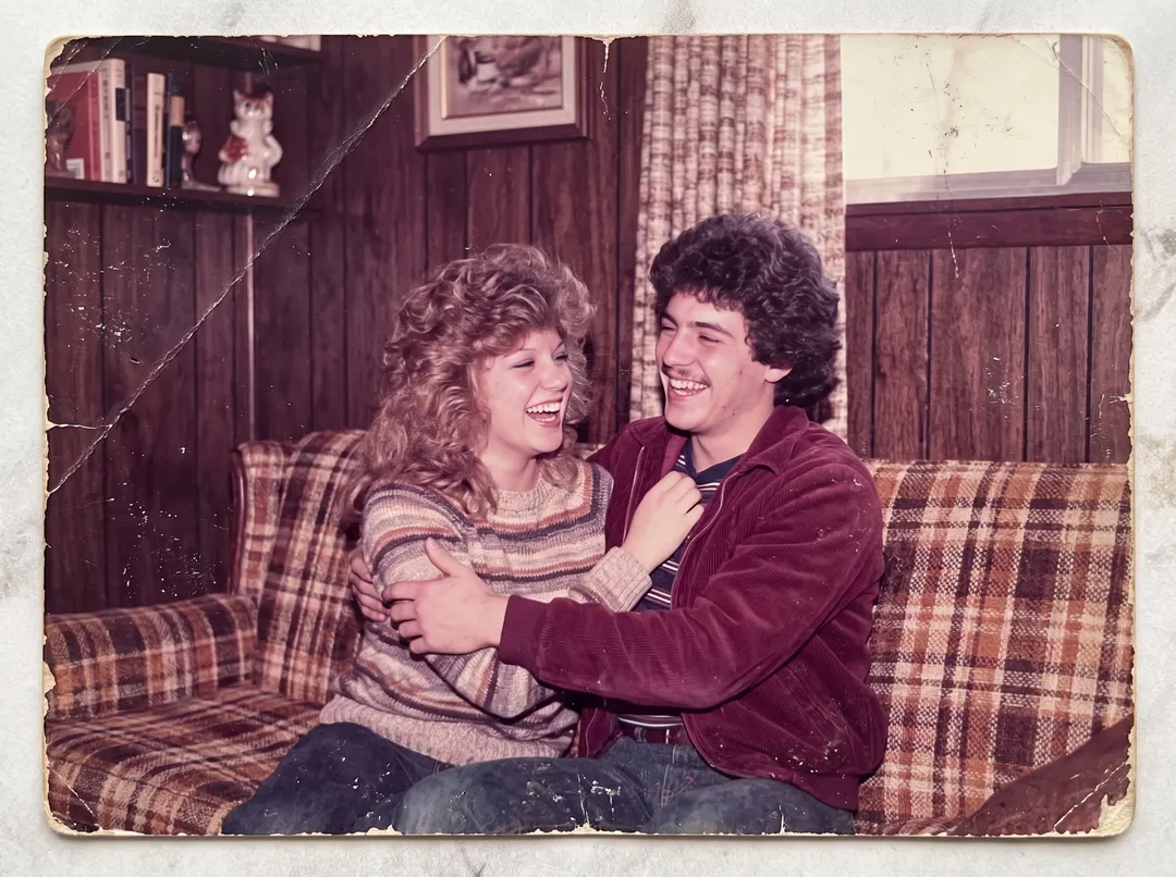 Damaged old photo of a 1980s couple laughing on a plaid sofa: magenta cast, scuffs, a crease