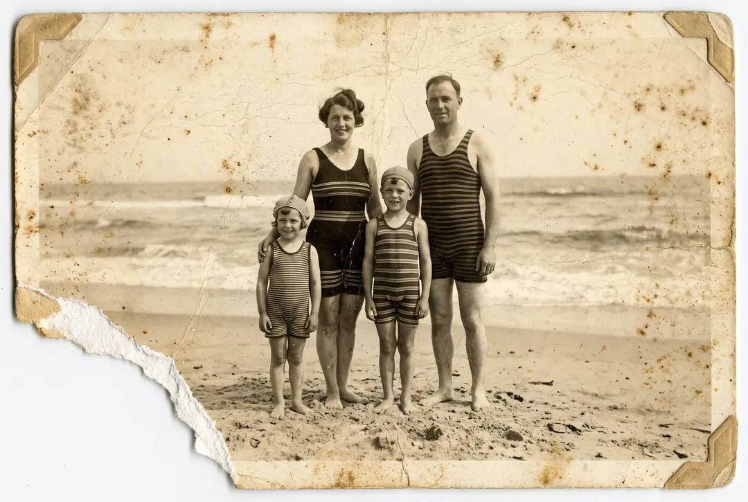 Damaged old photo of a 1930s family in striped bathing suits at the seaside: missing corner, foxing, cracked emulsion