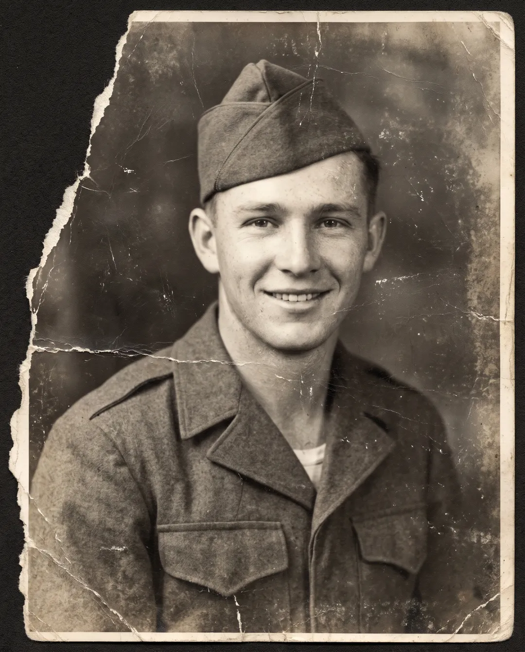Damaged old photo of a 1940s portrait of a young soldier in a garrison cap: missing strip, scratches, a crease