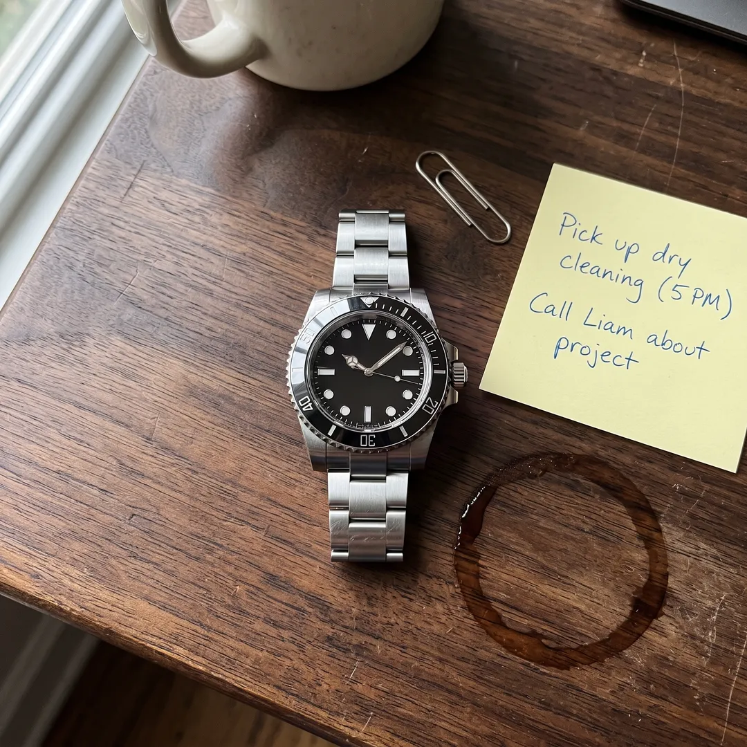 Silver dive watch on a walnut desk, with sticky note, paperclip and coffee stain in the shot