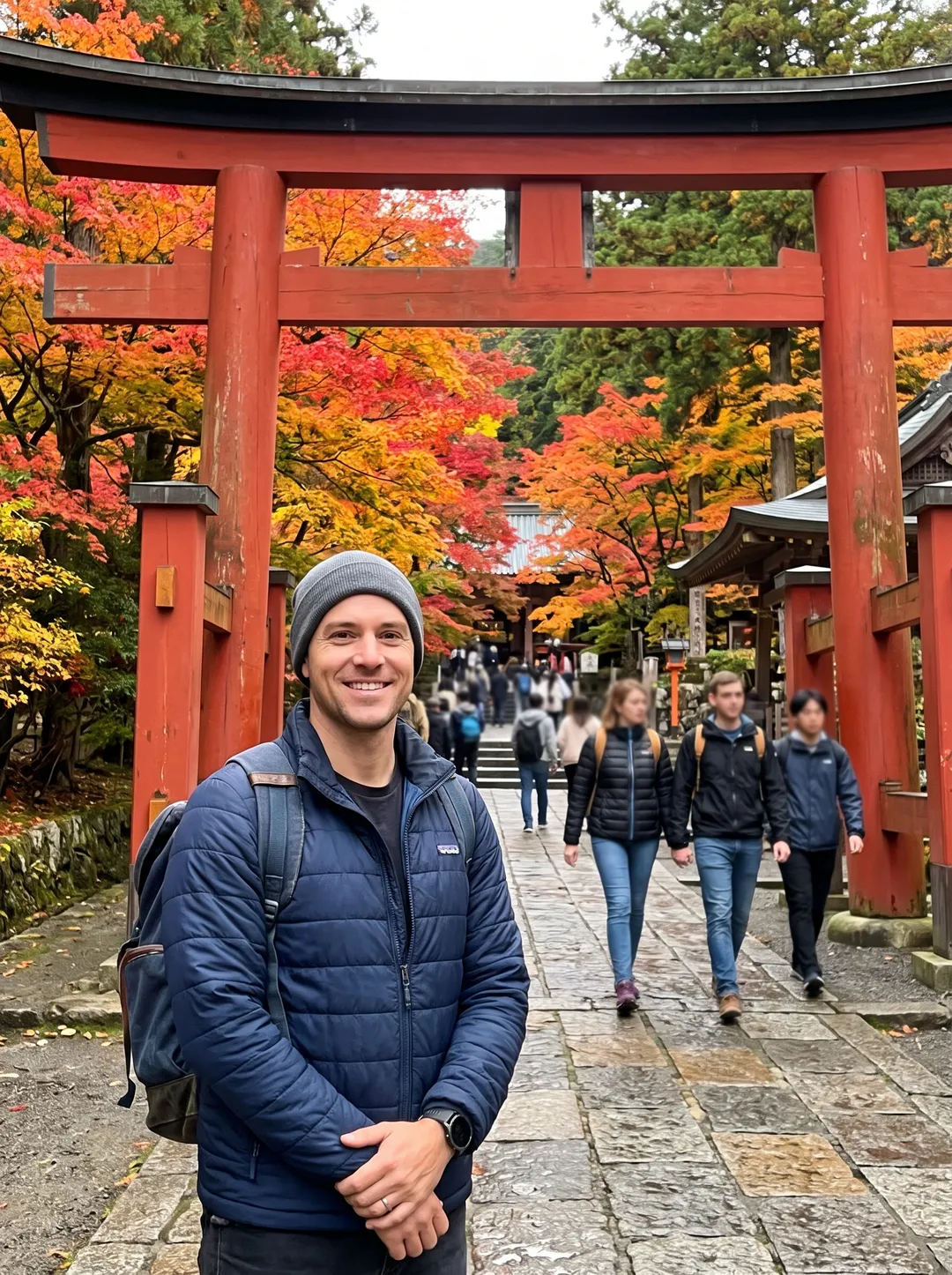 Photo of a man in a navy jacket in front of a red wooden temple gate in autumn, with other people in the frame
